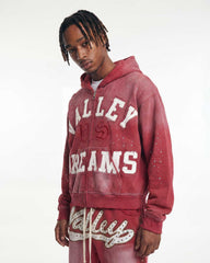 A man wearing a washed red zip-up hoodie with 'VALLEY DREAMS' printed in white on the front, paired with matching red sweatpants, against a plain white studio background.